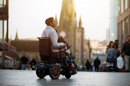 Brown skinned female powerchair user is shown driving her Q100 Powerchair with red trim in an urban setting. 