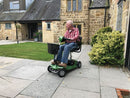 A man sits on a green mobility scooter with a front basket, parked on a stone patio in front of a stone house with a lawn and bushes.