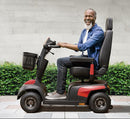 A man with a beard driving a compact electric mobility scooter on a paved walkway in front of a light gray stone wall and green hedges.