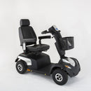 A modern black electric mobility scooter with a high-back seat, adjustable backrest, armrests, a front basket, and a small windshield, shown on a seamless light gray studio background.