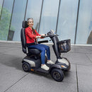 A woman wearing a red jacket and blue jeans is seated on a black and gray motorized mobility scooter with a front basket, riding on a paved walkway in front of a modern glass building.