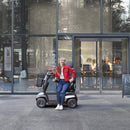 A woman wearing a red jacket, blue jeans, and white sneakers sits on a compact electric mobility scooter in front of a modern glass building.