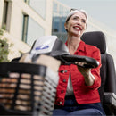 A smiling older woman with gray hair sits in a black motorized mobility chair or scooter seat, wearing a red button-up shirt and blue jeans, with a basket in the foreground and a modern building in the background.