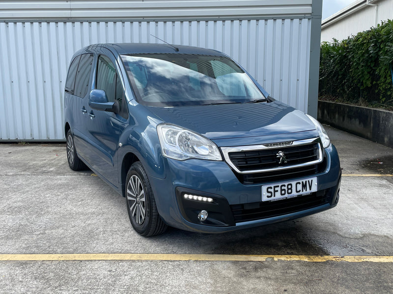 Blue Peugeot wheelchair accessible car parked on a concrete surface with a building and hedge in the background.