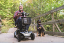 A man drives a Revo 2.0 Scooter across a wooden bridge in a woodland setting accompanied by his dog