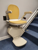 A reconditioned used approved straight stairlift with a beige chair and a footrest, situated at the top of a staircase.
